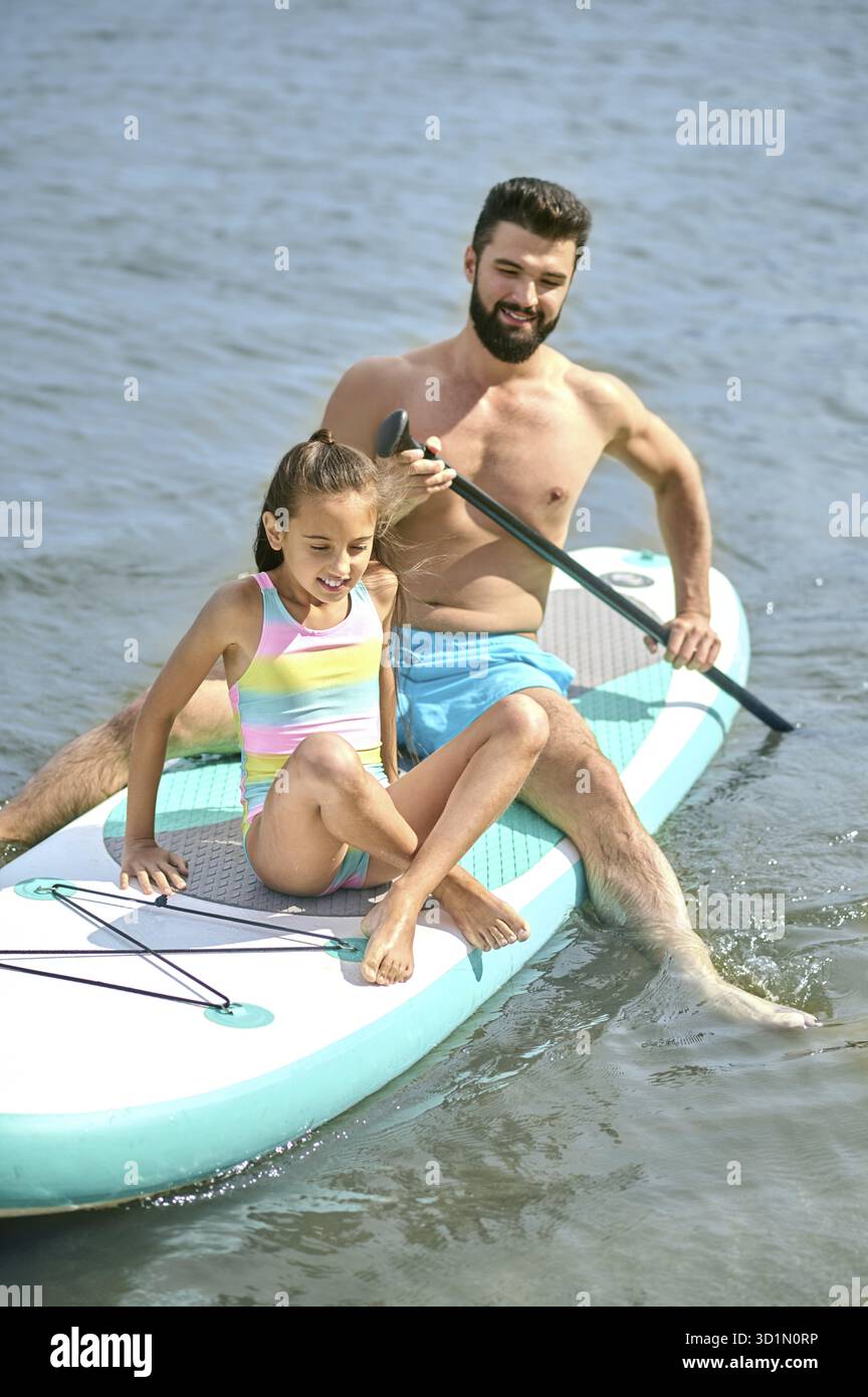 Atletico con sua figlia seduta in tavola da surf in acqua, godendo del tempo estivo insieme al resort Foto Stock