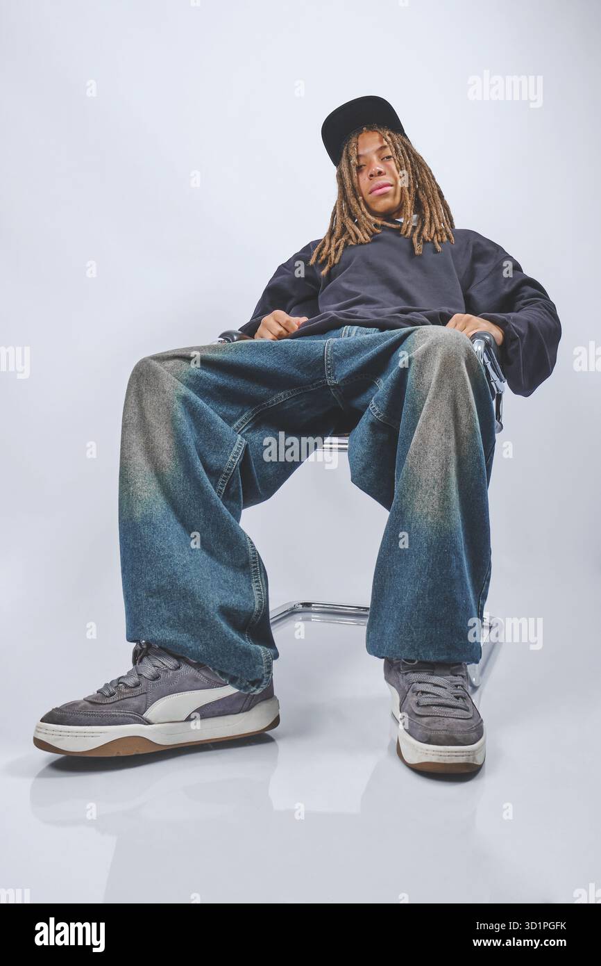 Giovane uomo con dreadlock e jeans larghi seduti su una sedia, che mette in risalto la moda urbana Foto Stock