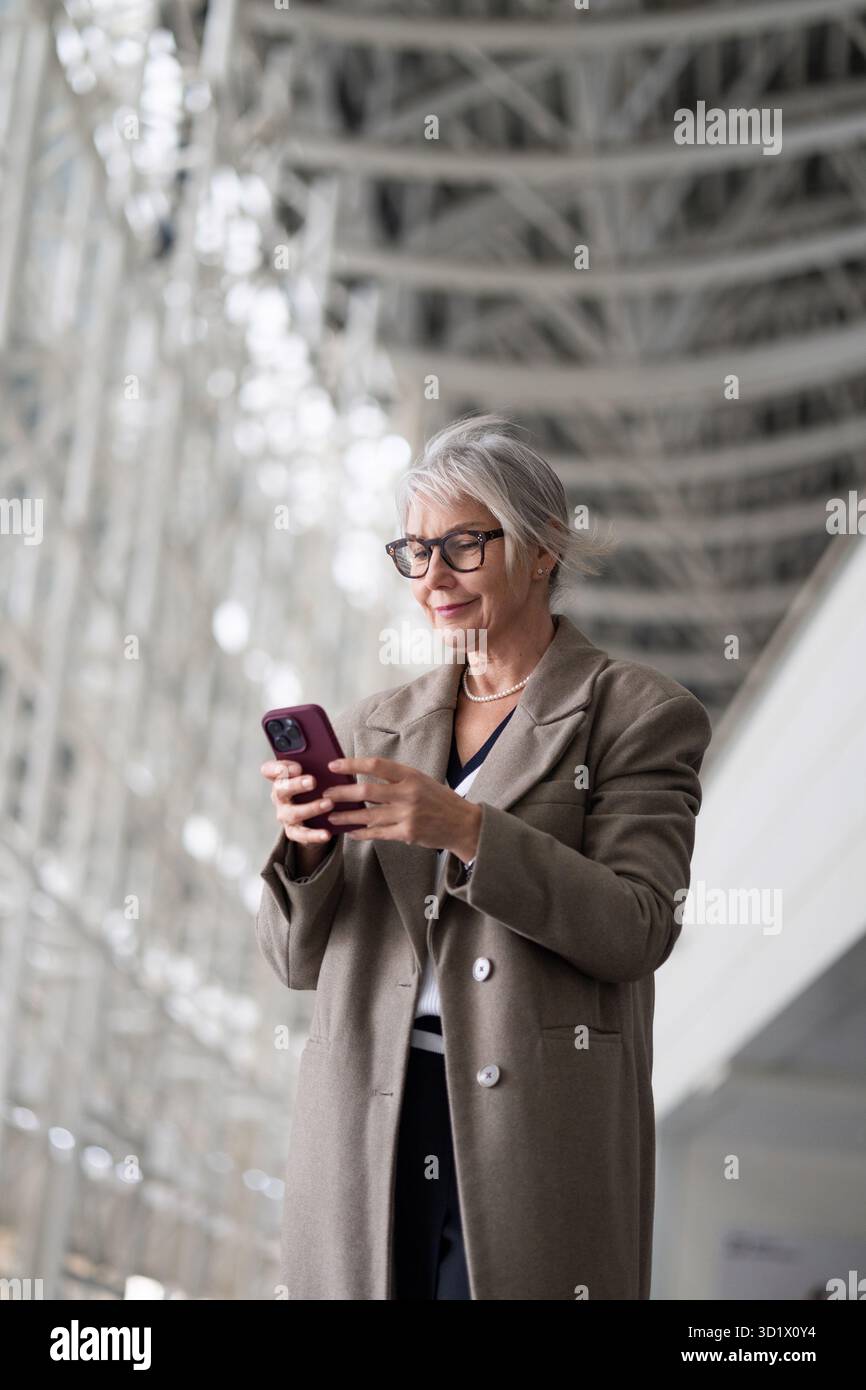 Elegante donna caucasica anziana che utilizza il suo smartphone mentre si trova in un ambiente architettonico moderno Foto Stock