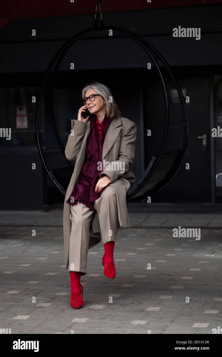Una donna anziana che si gode una conversazione al telefono mentre è seduta in un'altalena circolare in un moderno ambiente urbano all'aperto Foto Stock