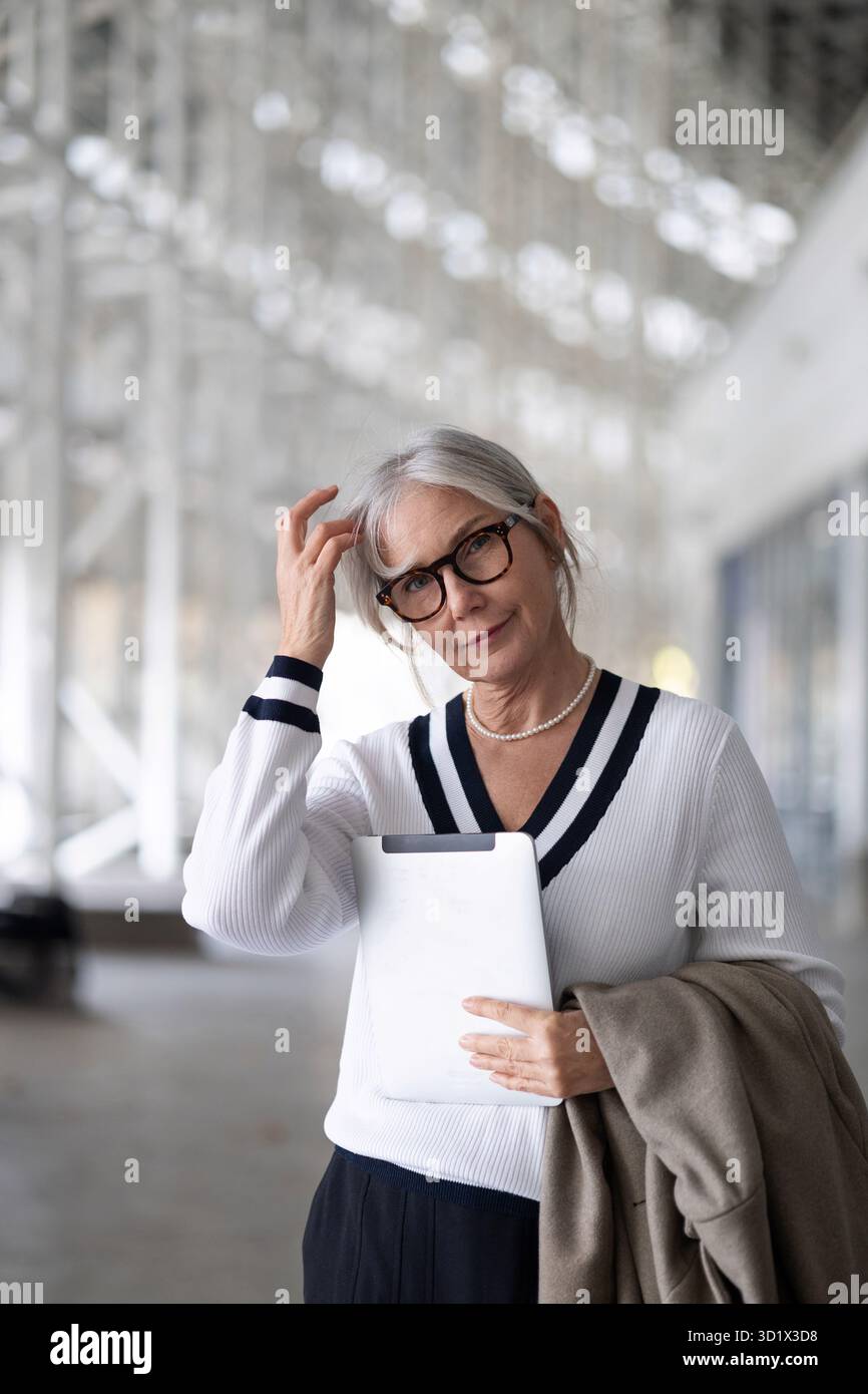 Una donna d'affari di mezza età riflette attentamente mentre tiene in mano un tablet in un ufficio moderno durante il pomeriggio Foto Stock
