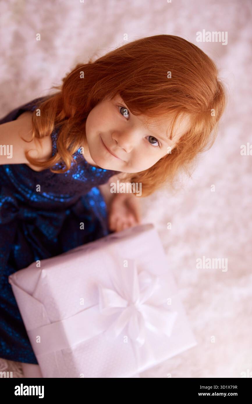 Gioioso bambino dalla testa rossa che tiene in mano una confezione regalo in un'atmosfera festosa con bianco Foto Stock