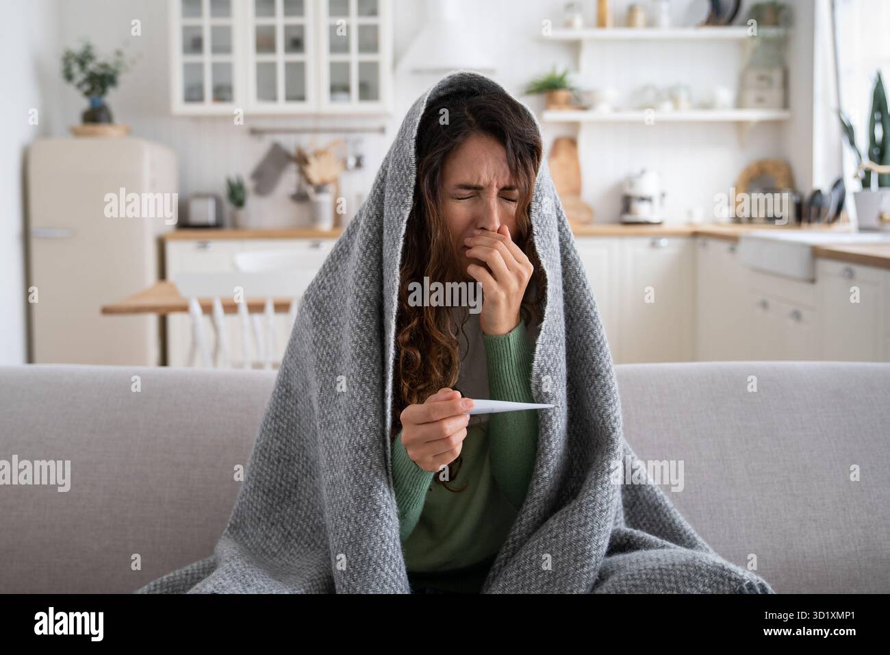 Donna malata seduta sul divano a casa con termometro nelle mani che soffre di naso che cola, starnuti Foto Stock