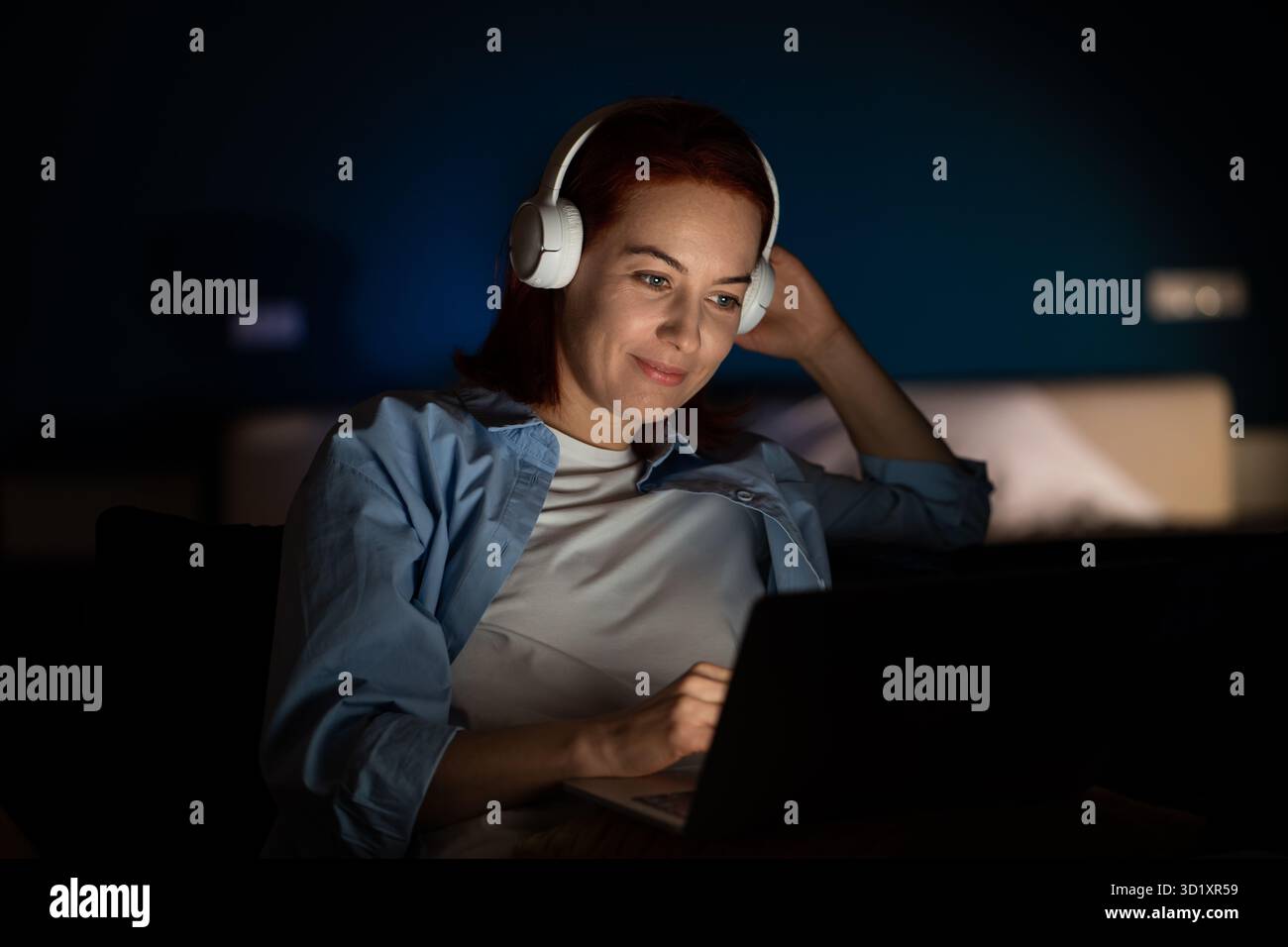 Donna calma con notebook guarda video con le cuffie, passa del tempo online a tarda notte in una stanza buia Foto Stock
