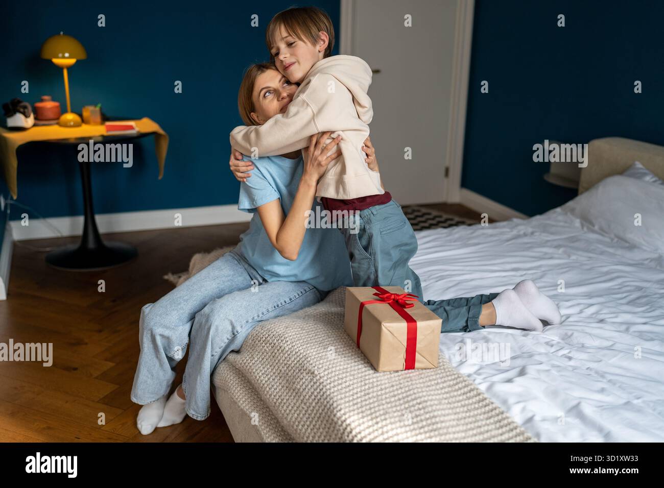 Mamma che dà regalo sorpresa il compleanno del figlio seduto a letto a casa, ragazzo abbracciando la donna. Foto Stock
