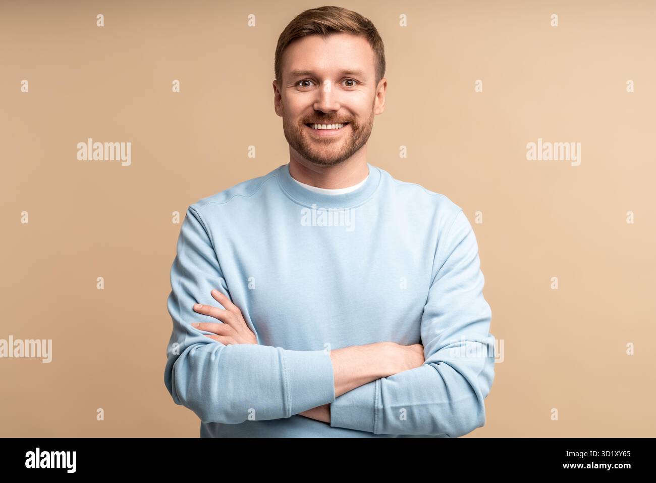Ritratto di un uomo sorridente con le mani incrociate sul petto su sfondo beige che guarda la fotocamera. Foto Stock