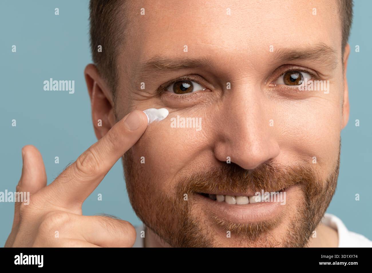 Bell'uomo che applica il prodotto di lozione per la pelle sul viso. Uomo attraente con pelle perfetta, panna cosmetica sorridente su sfondo blu. L Foto Stock