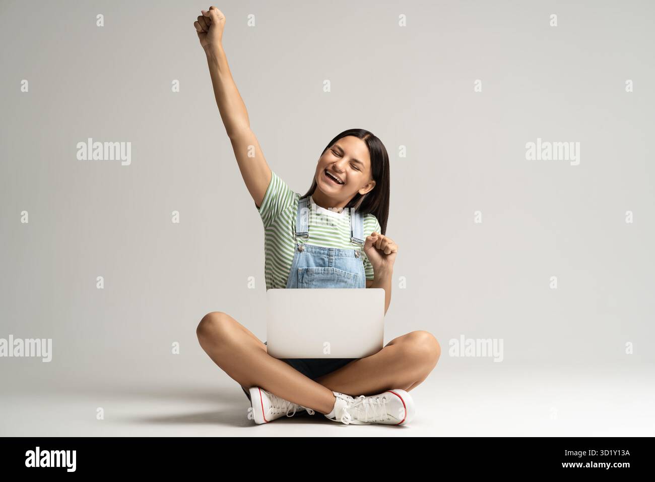 Felice ragazza adolescente sorridente che rallegra seduta su sfondo grigio con il laptop sulle gambe. Foto Stock