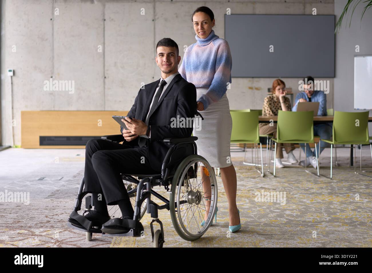 In un ufficio professionale, una donna d'affari che aiuta il suo direttore, seduto su una sedia a rotelle e che utilizza un tablet, mentre è in viaggio Foto Stock