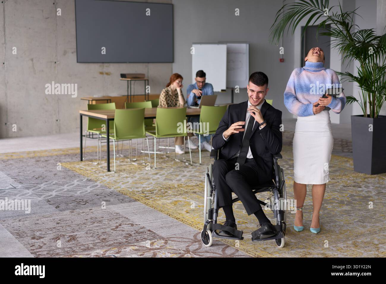 In un ufficio professionale, una donna d'affari che aiuta il suo direttore, seduto su una sedia a rotelle e che utilizza un tablet, mentre è in viaggio Foto Stock