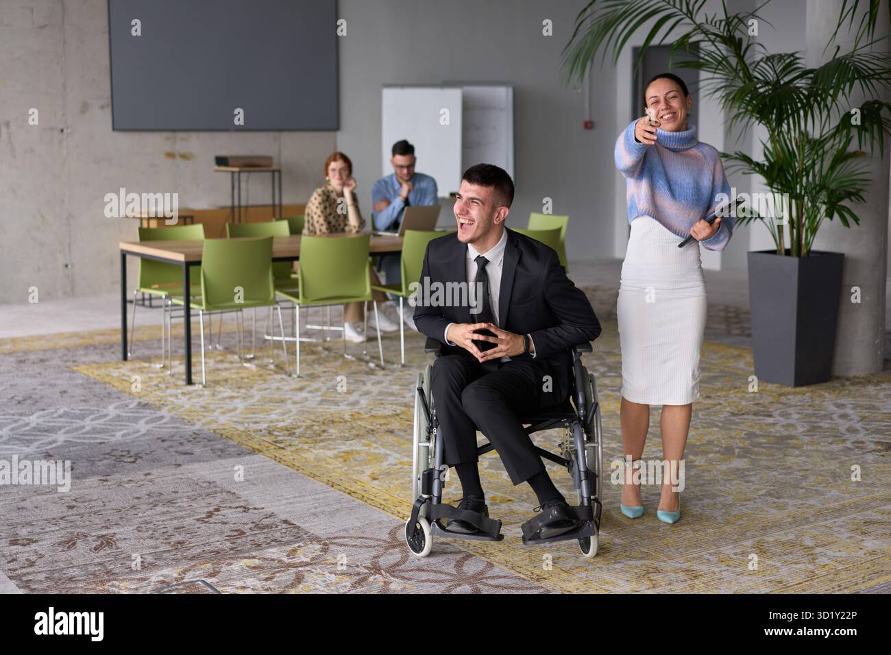 In un ufficio professionale, una donna d'affari che aiuta il suo direttore, seduto su una sedia a rotelle e che utilizza un tablet, mentre è in viaggio Foto Stock