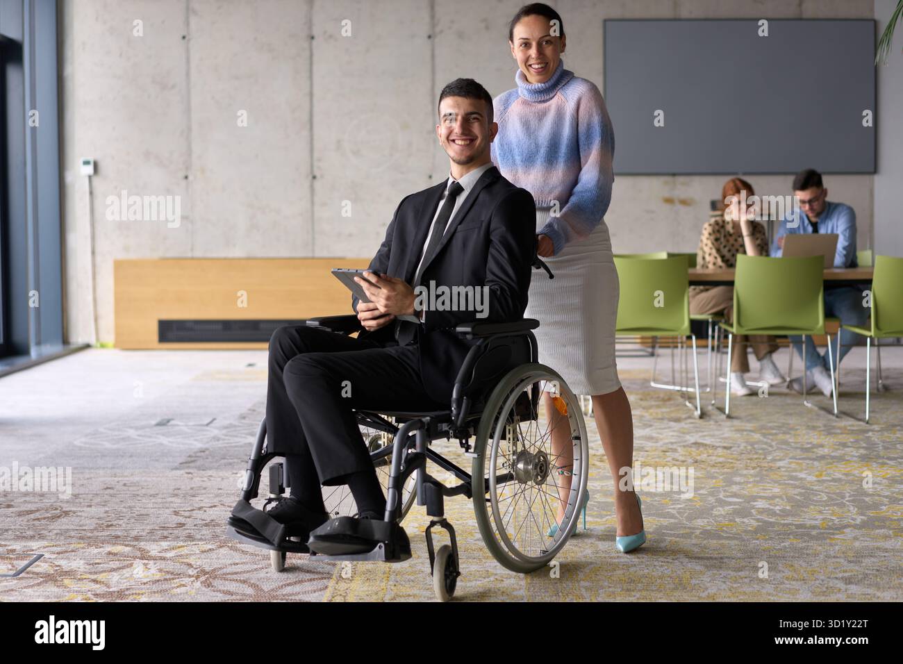 In un ufficio professionale, una donna d'affari che aiuta il suo direttore, seduto su una sedia a rotelle e che utilizza un tablet, mentre è in viaggio Foto Stock