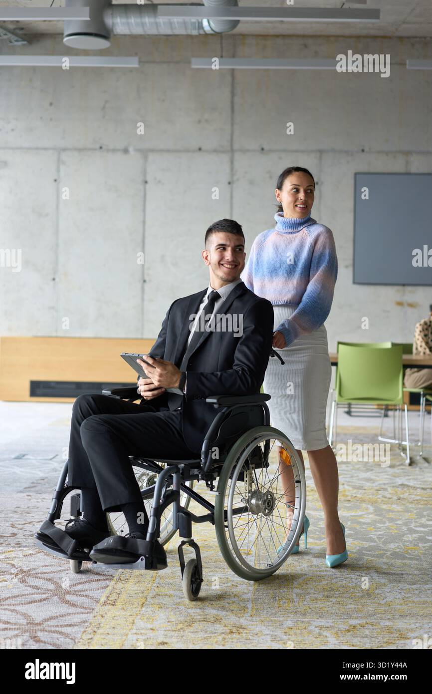 In un ufficio professionale, una donna d'affari che aiuta il suo direttore, seduto su una sedia a rotelle e che utilizza un tablet, mentre è in viaggio Foto Stock