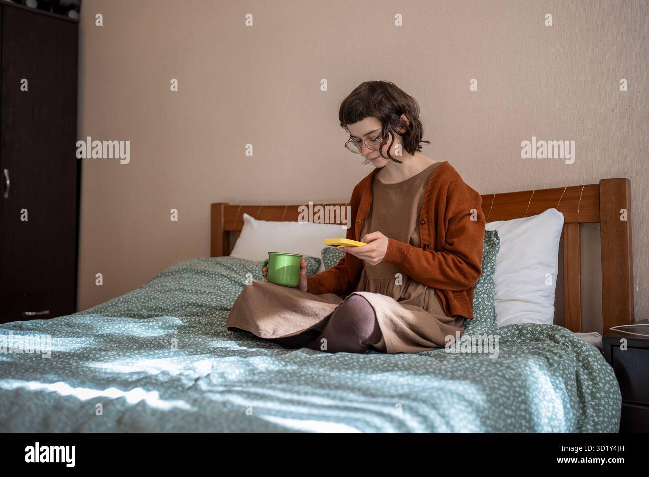 Ragazza adolescente che legge messaggi scorrendo i social media nello smartphone bevendo tè a letto. Dipendenza telefonica Foto Stock