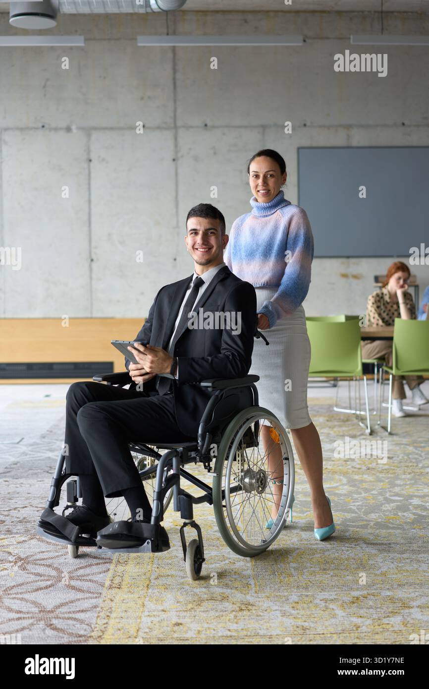 In un ufficio professionale, una donna d'affari che aiuta il suo direttore, seduto su una sedia a rotelle e che utilizza un tablet, mentre è in viaggio Foto Stock