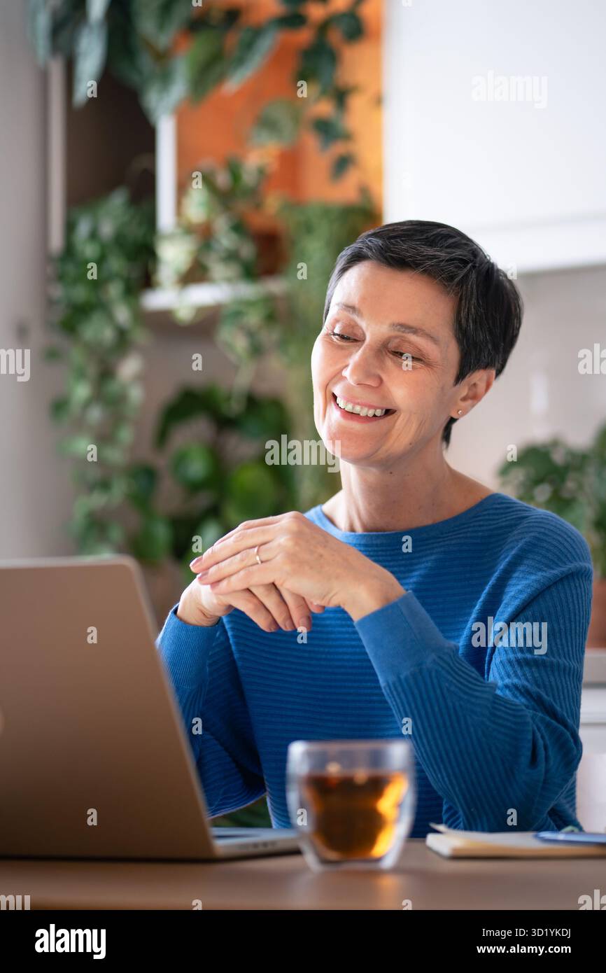 Una donna sorridente sorridente si diverte a gustare il tè mentre effettua videochiamate sul laptop a casa. Apprendimento online, incontro virtuale Foto Stock