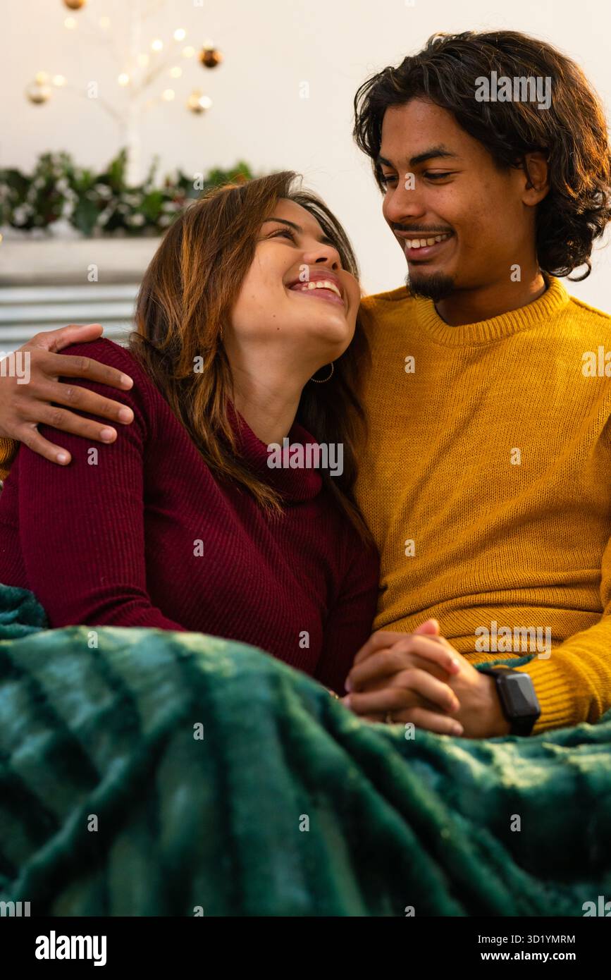 Coppie diverse che si accoccolano sotto la coperta verde sul divano accanto al camino con ghirlande e baubles Foto Stock