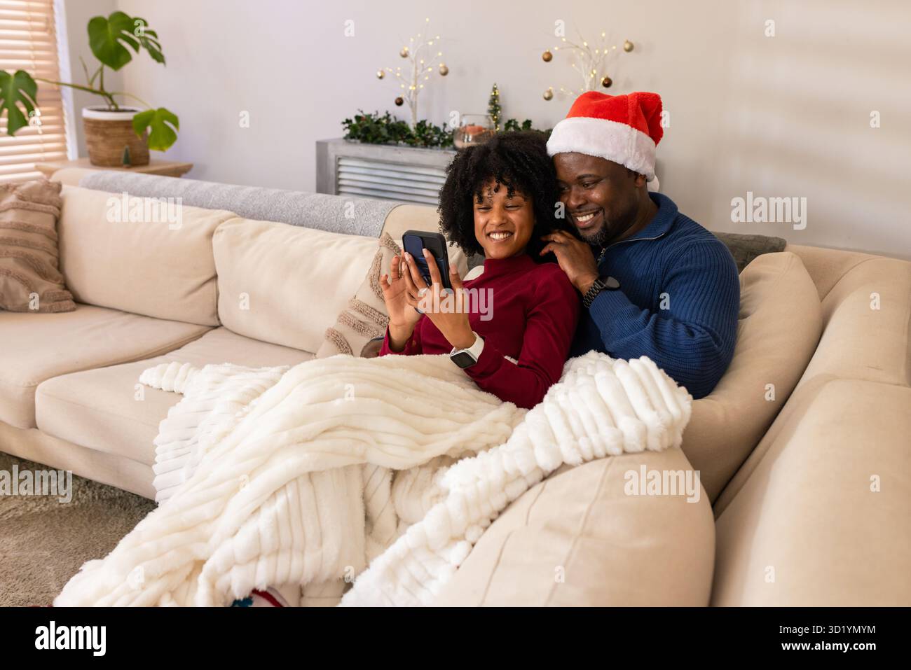 Coppia afroamericana che si coccola sotto la coperta sul divano di casa guardando lo smartphone vicino all'albero delle vacanze Foto Stock
