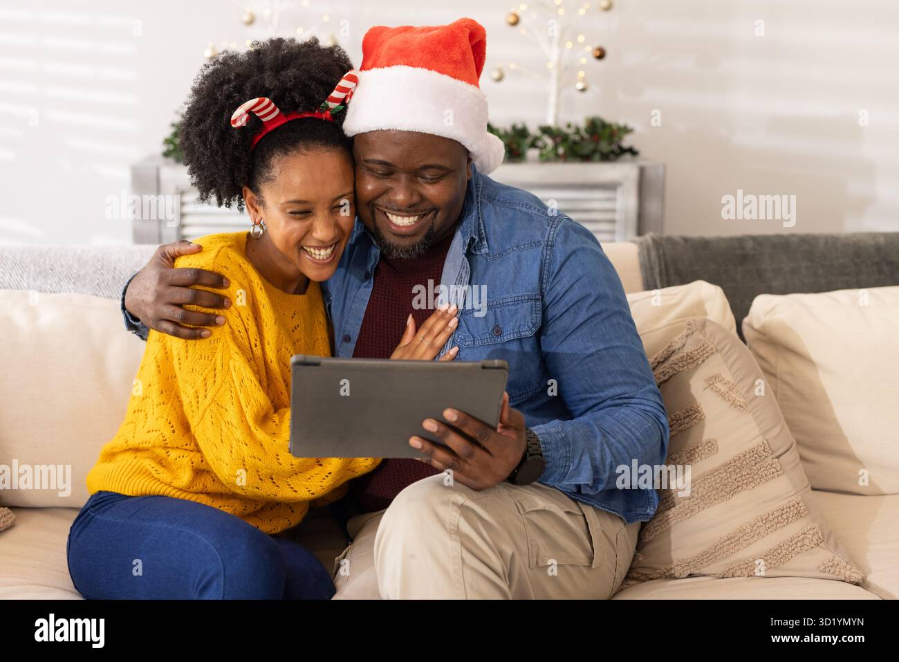 Coppia variegata che indossa il cappello di Babbo Natale e l'archetto di canna da zucchero che si coccola mentre guarda il tablet sul divano Foto Stock