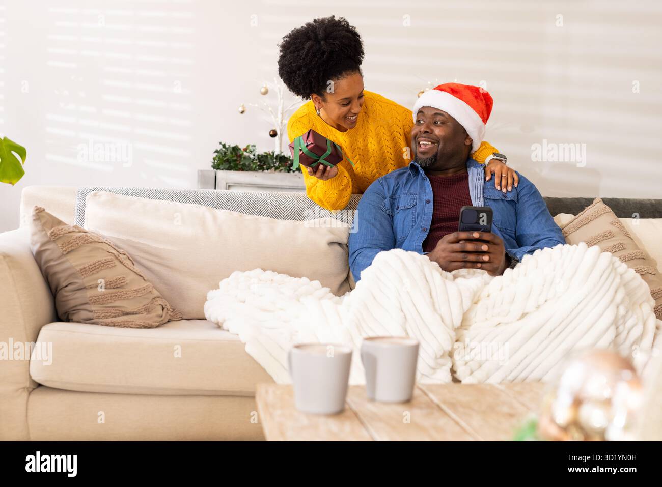Coppia afroamericana che si scambia il regalo di Natale seduto sul divano con lo smartphone a casa Foto Stock