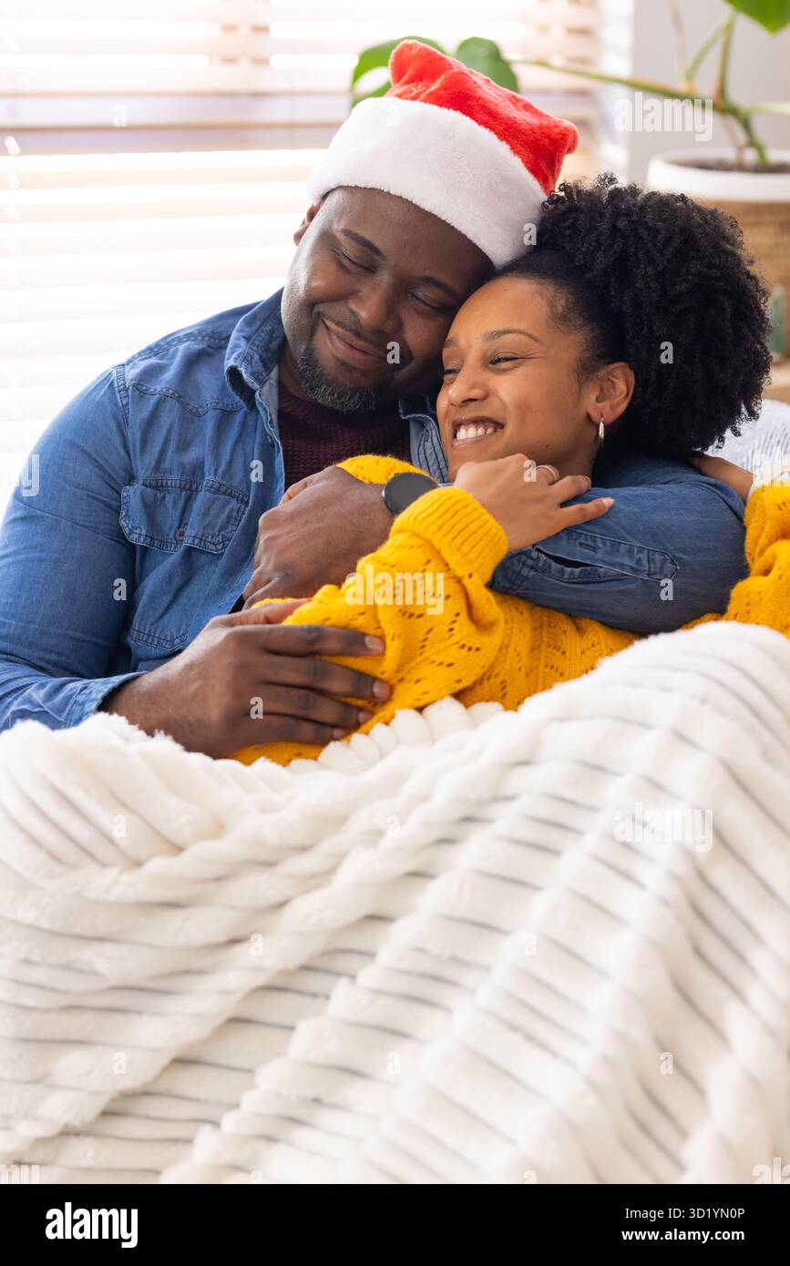 Coppia afroamericana che indossa il cappello di Babbo Natale coccolandosi sotto una coperta bianca sul divano nel soggiorno Foto Stock