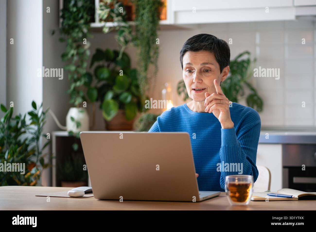 Gesti di una donna d'affari matura e impegnata che spiegano le attività di una riunione video a casa con tè e note Foto Stock