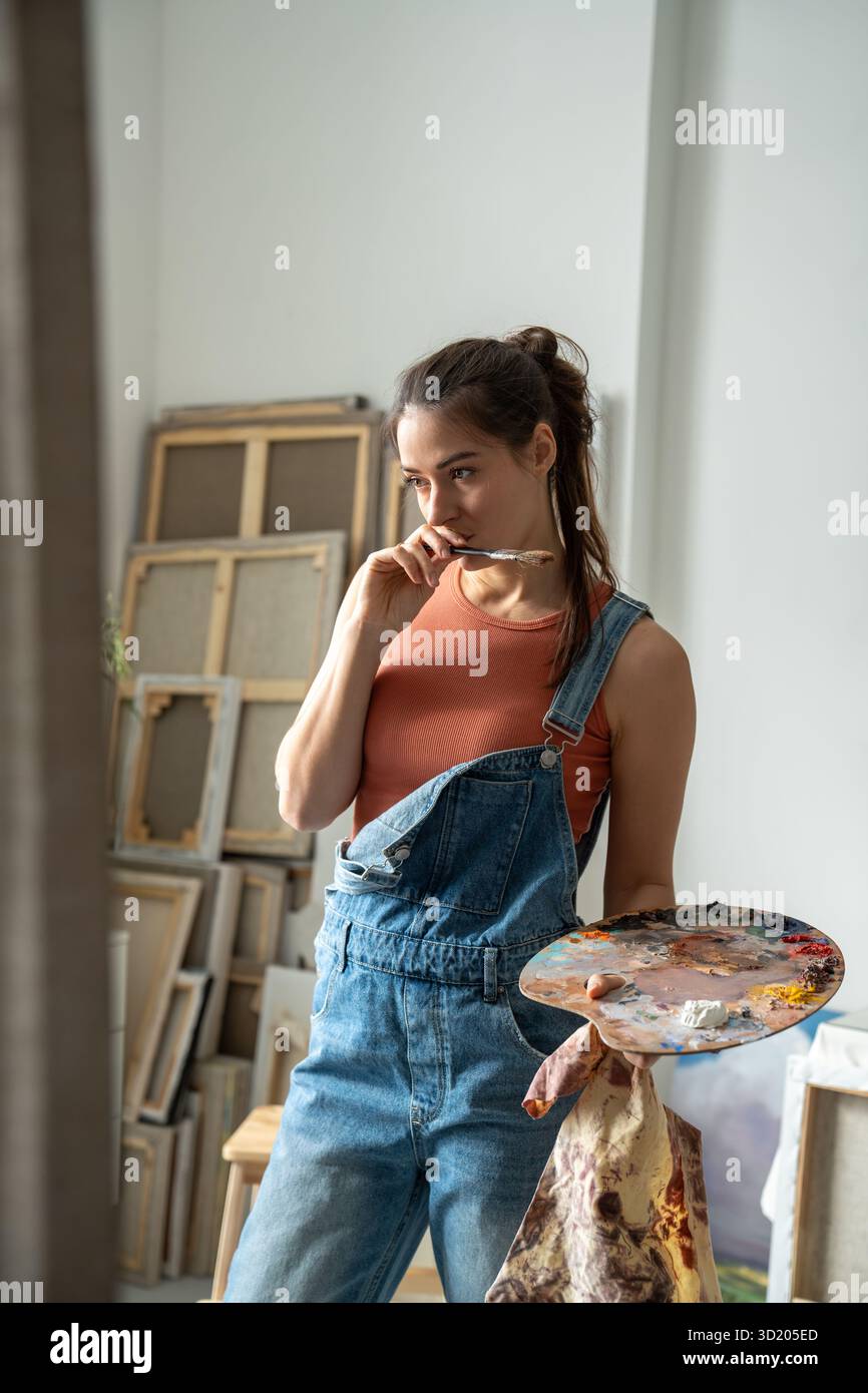 Una donna ispirata ha una tavolozza, un pensivo look da dipingere su tela, pensa all'idea in uno studio d'arte Foto Stock