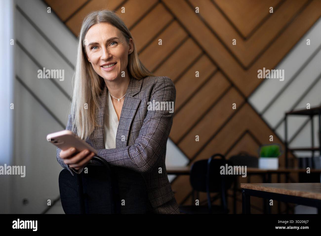 Donna professionista in un moderno ambiente di ufficio che utilizza lo smartphone seduto su una sedia con elegante sfondo in legno Foto Stock