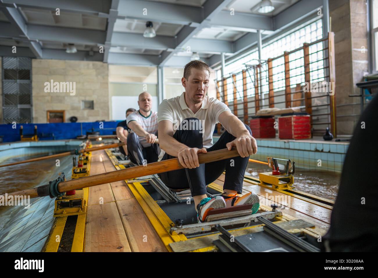 Gli atleti affaticati si allenano rigorosamente sul simulatore di canottaggio nella piscina della palestra superando lo sfinimento Foto Stock