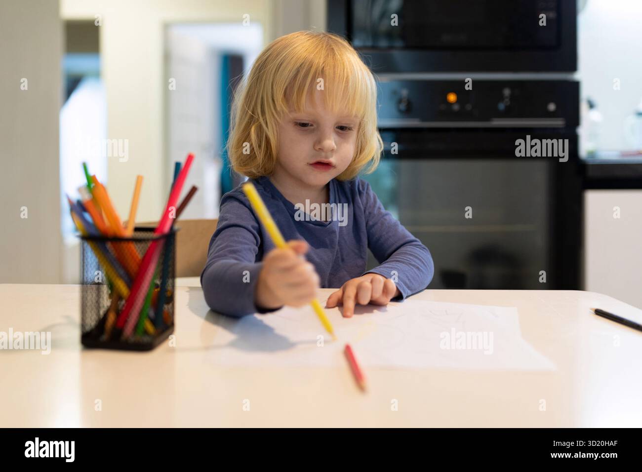 Il bambino si impegna nel disegno creativo al tavolo della cucina utilizzando pennarelli colorati durante le ore diurne Foto Stock