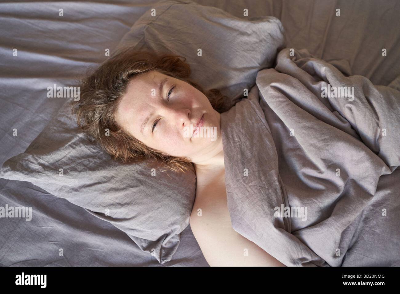 Donna caucasica adulta che si rilassa nel letto con comodi letti grigi e luce naturale Foto Stock