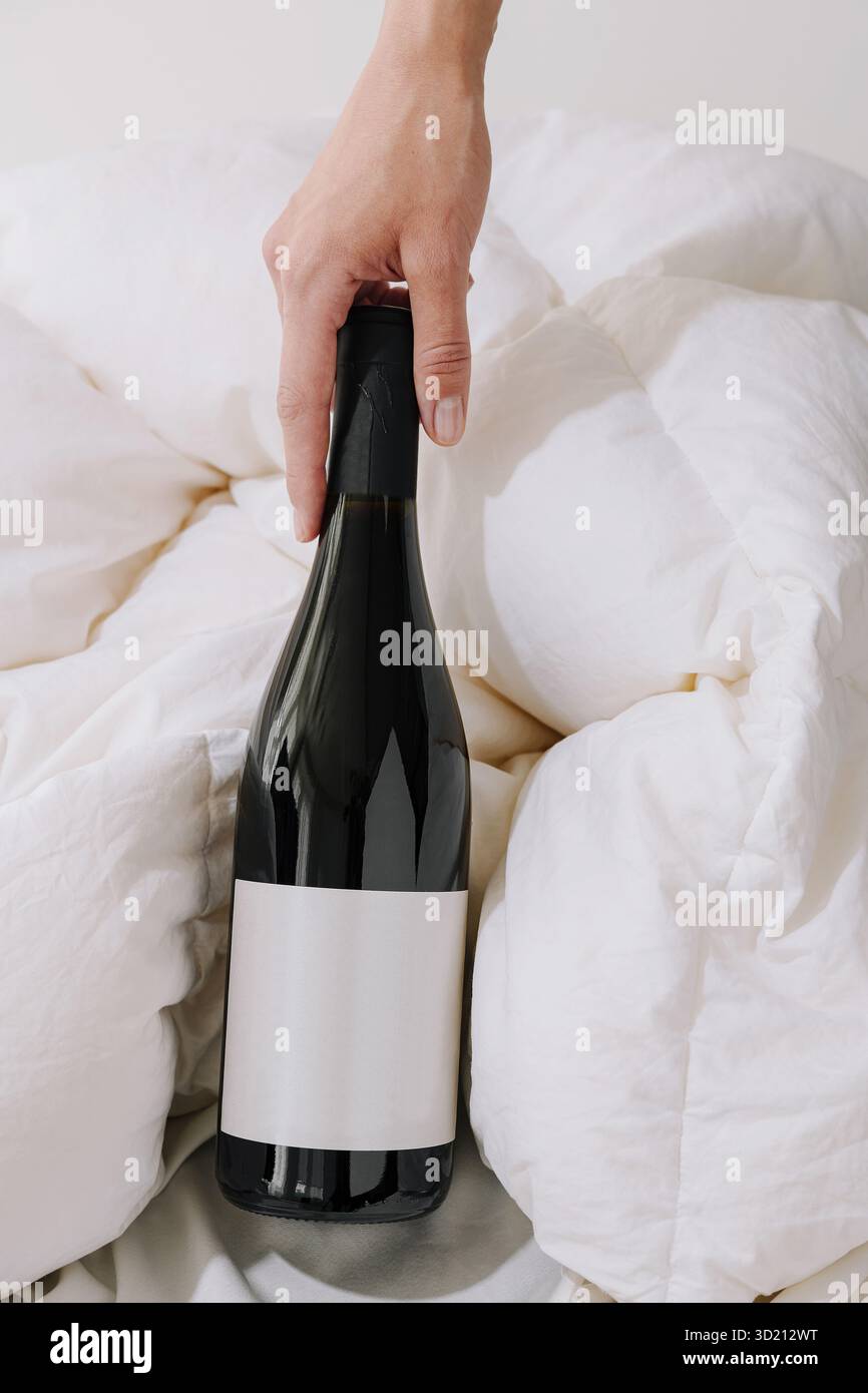 La mano arriva per una bottiglia di vino nero appoggiata su una coperta bianca in un ambiente accogliente durante il giorno Foto Stock