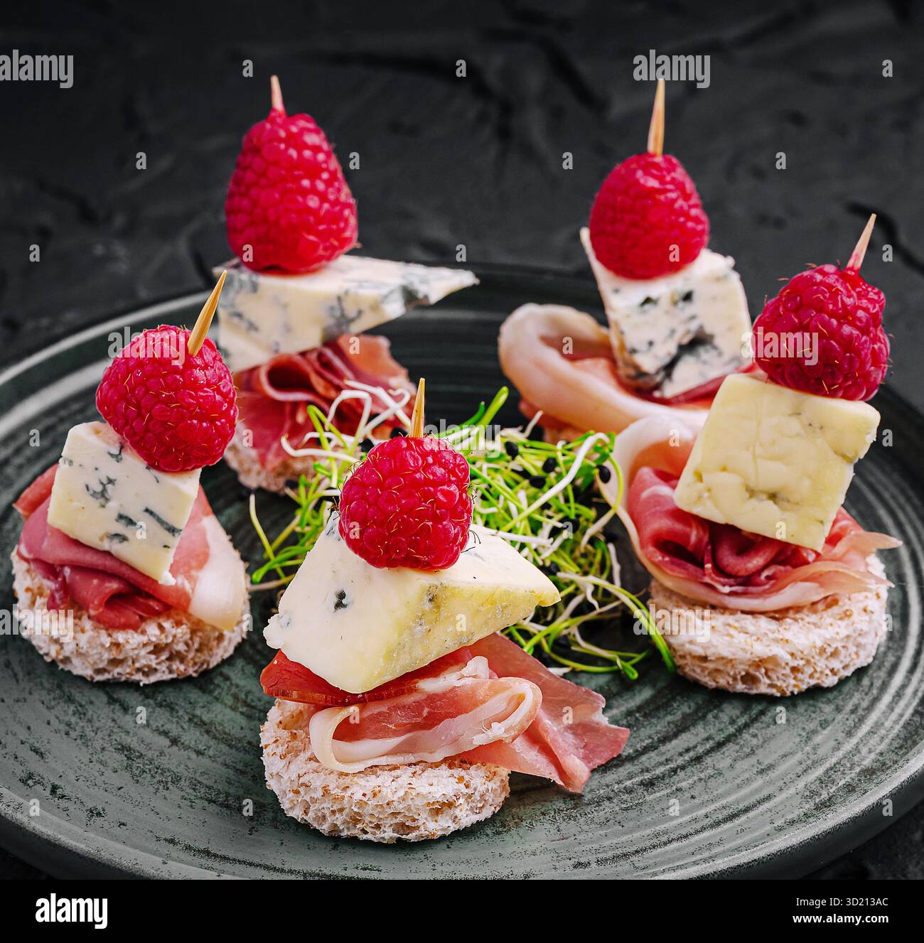 Deliziosi piccoli bocconcini con strati di formaggio, prosciutto e lamponi freschi su un piatto scuro. Perfetto per riunioni o cene di lusso, offe Foto Stock
