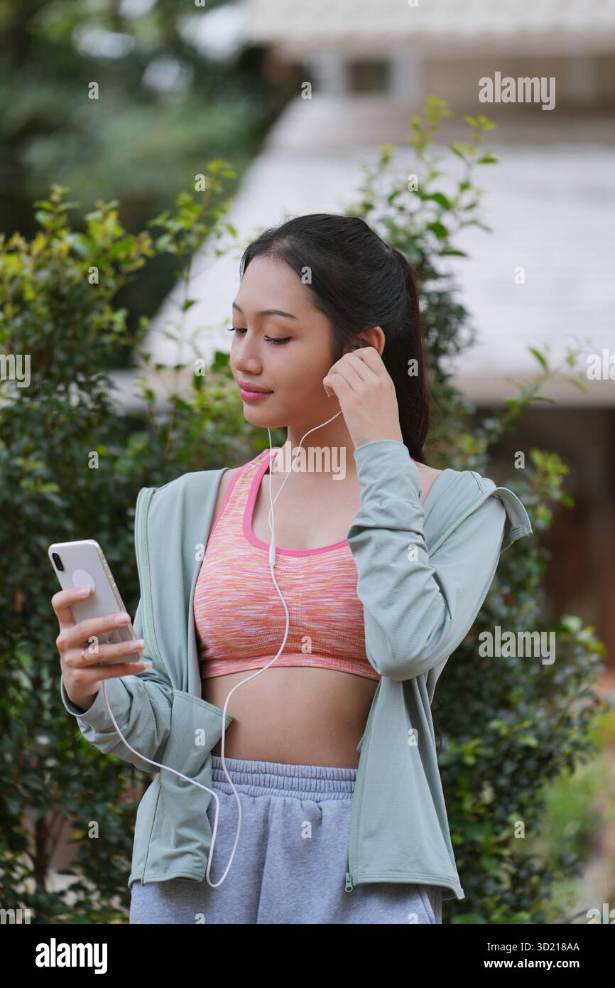 Adatta la giovane donna asiatica utilizzando il telefono che controlla le applicazioni di fitness in piedi nel verde del parco naturale all'aperto. Foto Stock