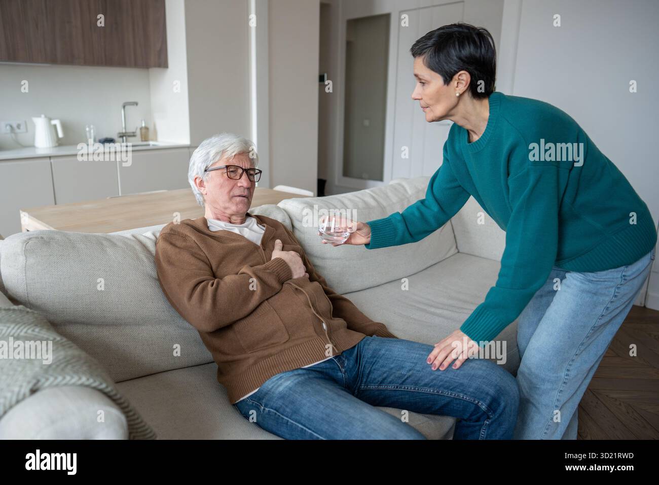 La moglie premurosa aiuta gli anziani che soffrono marito a stringere il petto seduto sul divano, offrendo un bicchiere d'acqua Foto Stock