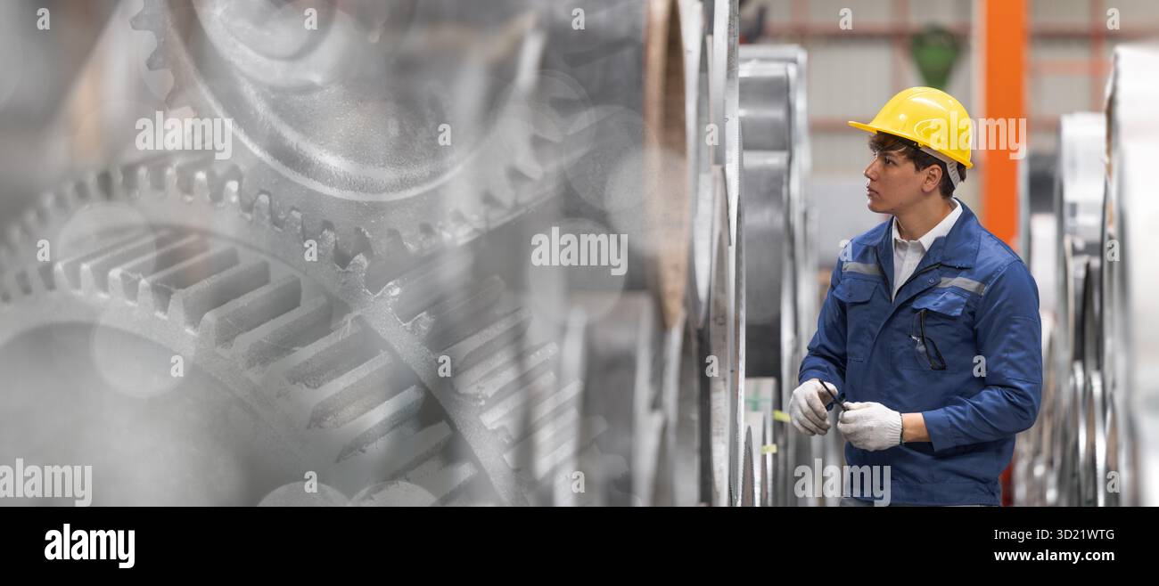 Operaio industriale con elmetto di sicurezza blu e giallo che ispeziona macchinari in fabbrica, il monitoraggio tecnico e gli ingranaggi hanno un background ampio f Foto Stock