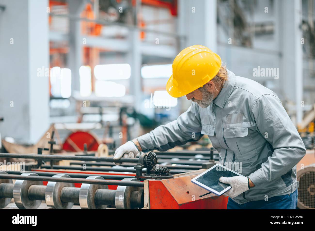 Lavoratore industriale senior professionista in caschi protettivi e macchinari per l'assistenza uniforme in fabbrica, tecnico esperto addetto alla manutenzione di macchinari pesanti Foto Stock