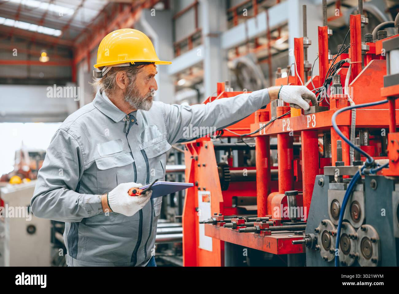 Lavoratore industriale senior professionista in caschi protettivi e macchinari per l'assistenza uniforme in fabbrica, tecnico esperto addetto alla manutenzione di macchinari pesanti Foto Stock