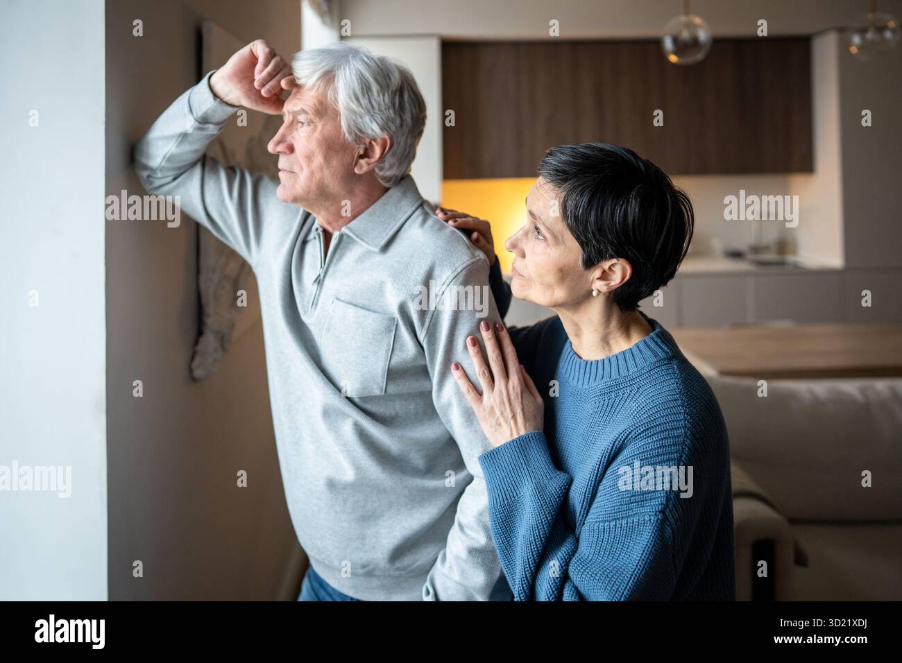 Un uomo anziano triste e premuroso guarda fuori dalla finestra mentre la moglie si allontana, consolando. Supporto familiare, empatia Foto Stock