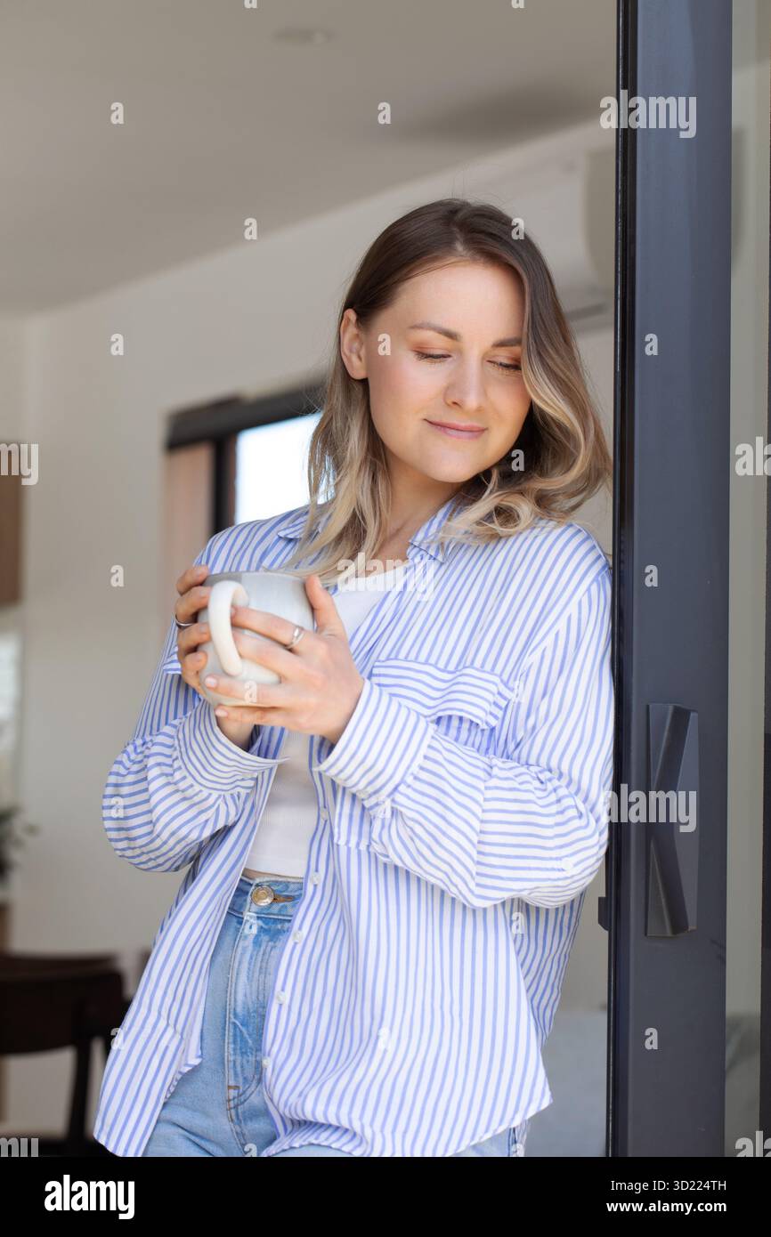 Donna positiva in camicia a righe tiene una tazza di caffè in terrazza Foto Stock