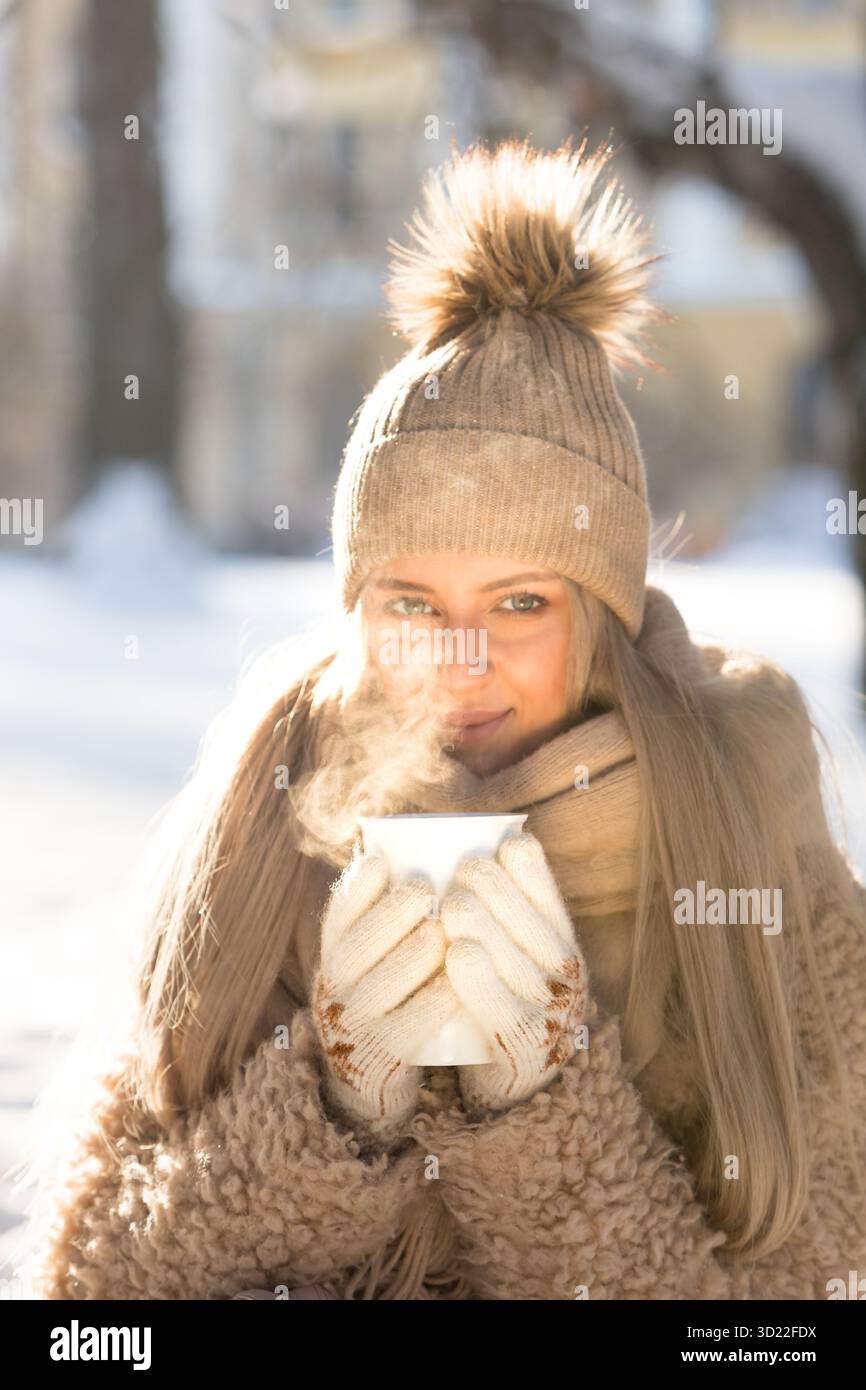 Donna che tiene in mano una tazza di tè o caffè caldo, all'aperto nella soleggiata giornata invernale guardando la macchina fotografica Foto Stock