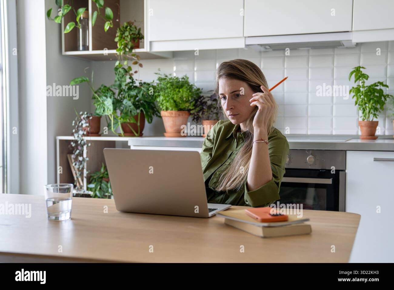 Donna stanca e premurosa che lavora su un laptop, lezioni virtuali online circondate da libri e bicchieri d'acqua Foto Stock