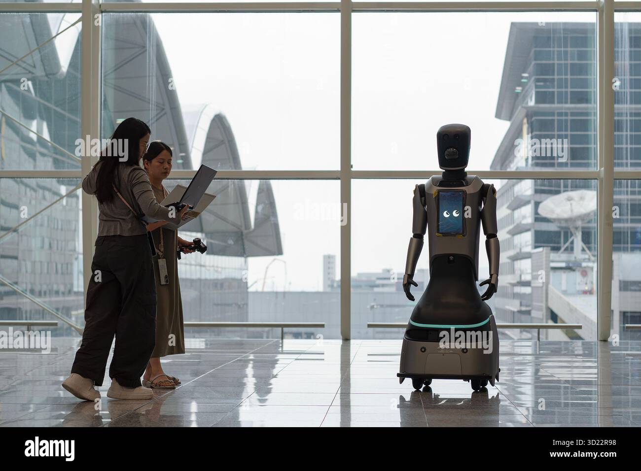 Hong Kong, Cina - 20 settembre 2025: Due donne stanno in piedi e tengono in mano lapplani vicino a un robot di servizio nell'aeroporto internazionale di Hong Kong. Foto Stock