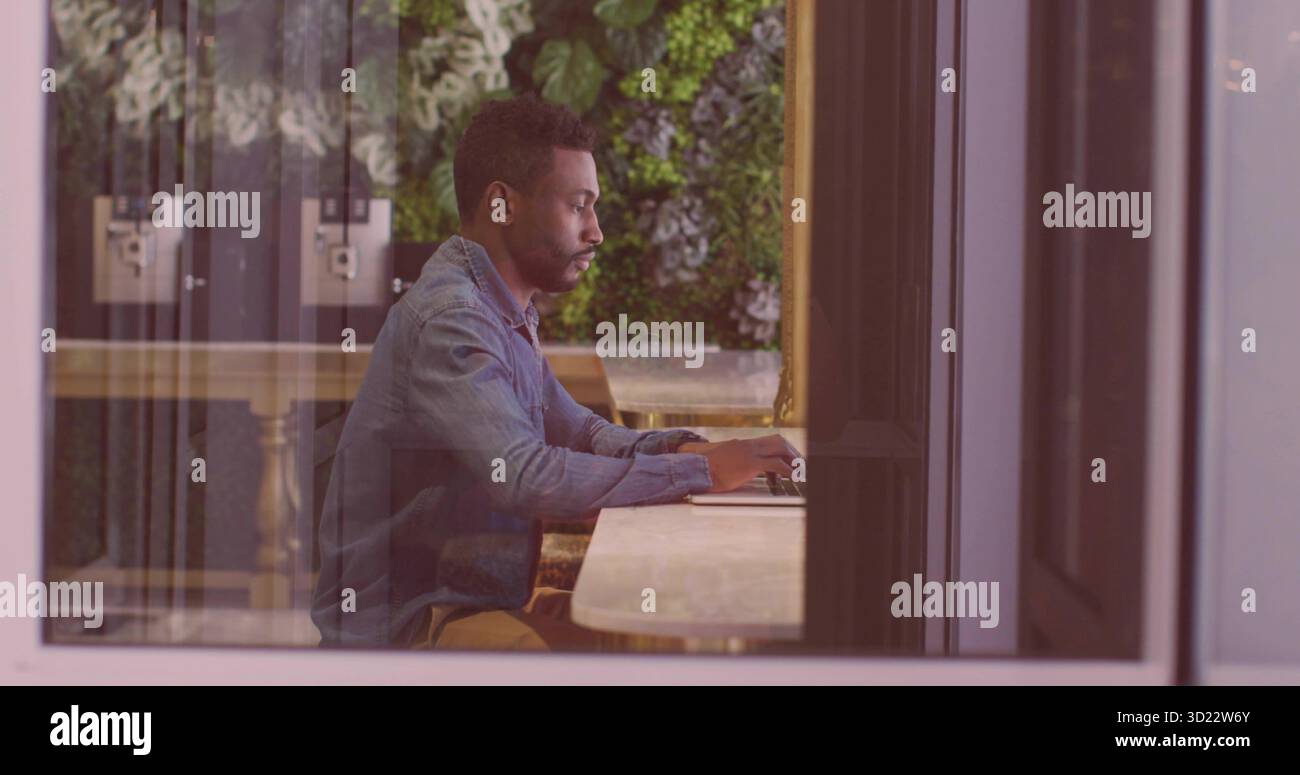 Uomo che scrive indossa camicia in denim al banco di marmo del caffè, con computer portatile, parete dello stabilimento e dispenser Foto Stock