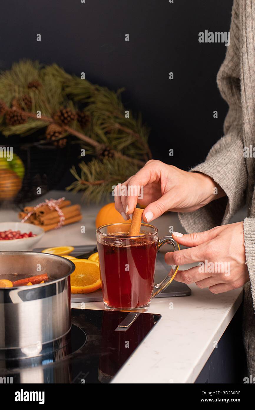 Una donna si trova vicino alla stufa con una pentola di VIN brulé e immerge un bastoncino di cannella in un bicchiere di VIN brulé caldo. Foto Stock