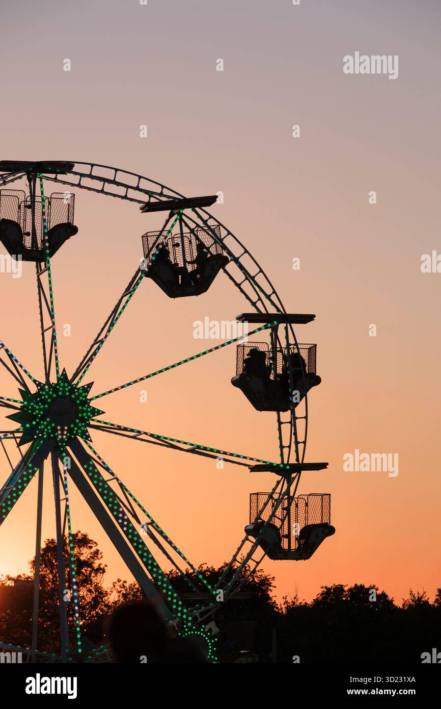 Silhouette di persone che cavalcano una ruota panoramica contro un cielo serale rosa/arancione Foto Stock