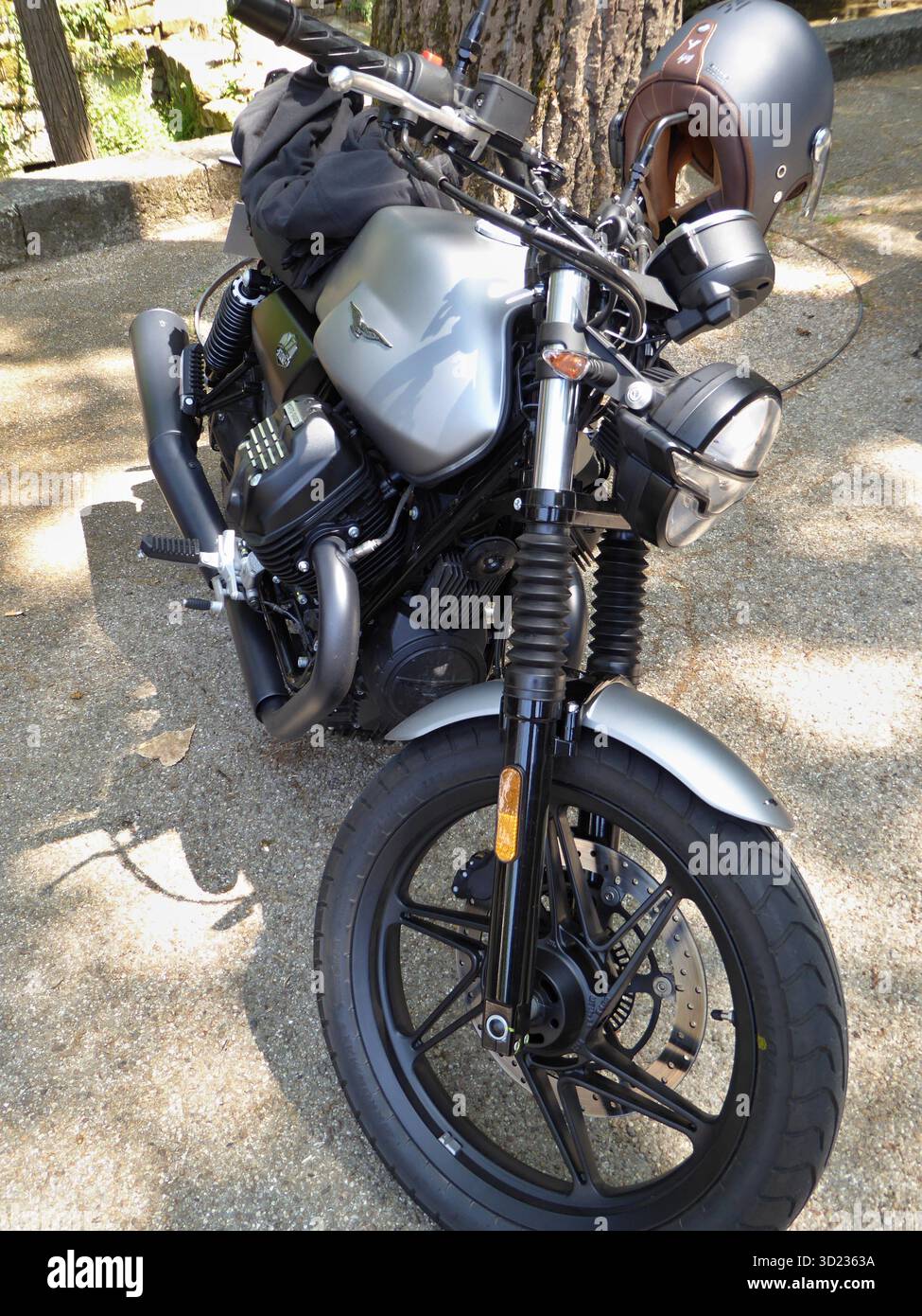Moto Guzzi V7 Stone con casco marrone appeso al manubrio parcheggiato all'ombra di un albero Foto Stock