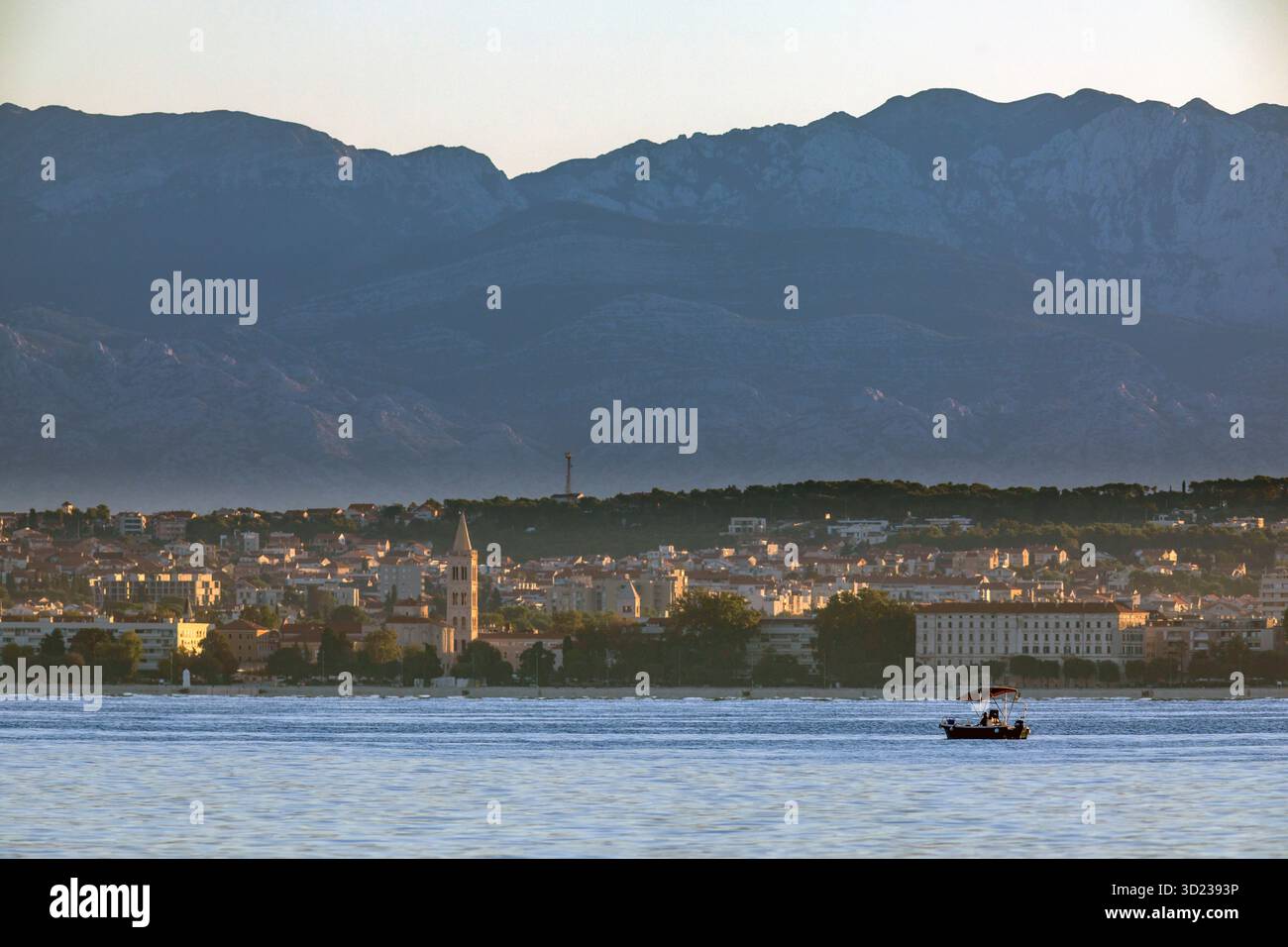 Una barca naviga sulle acque calme con la città di Zara e lo sfondo delle montagne sotto un cielo limpido, la Croazia. Foto Stock