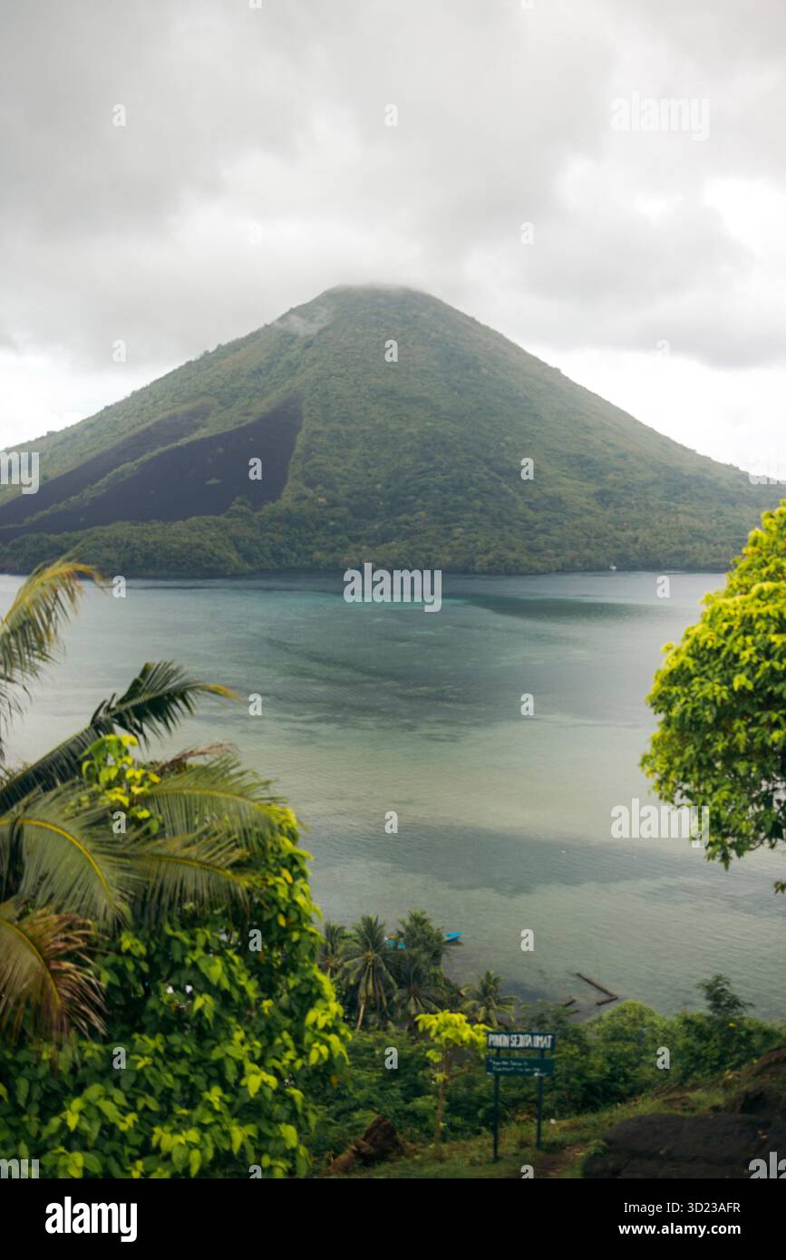 Lussureggiante montagna verde sorge su una baia tranquilla e nuvolosa con piante tropicali in primo piano. Banda Neira, banda, Maluku centrale, Indonesia Foto Stock