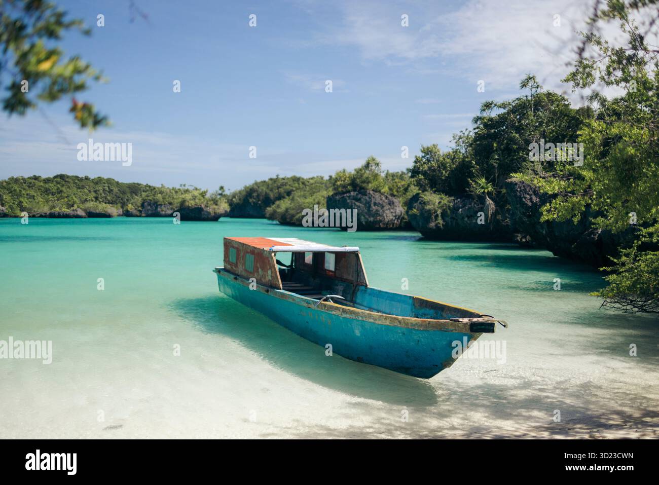 Una barca blu sulle acque turchesi vicino a una lussureggiante costa tropicale verde con luce solare intensa. Pulau Bair o Bair Island a Kei Kecil, Reggenza sud-orientale di Maluku, Maluku, Indonesia Foto Stock
